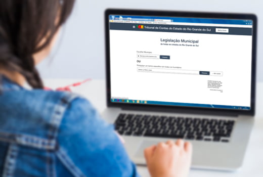 Audiodescrição: A imagem tem o fundo levemente desfocado, e mostra uma mulher usando um notebook. Ela está acessando a nova plataforma de Legislação Municipal do TCE-RS (fim da descrição).
